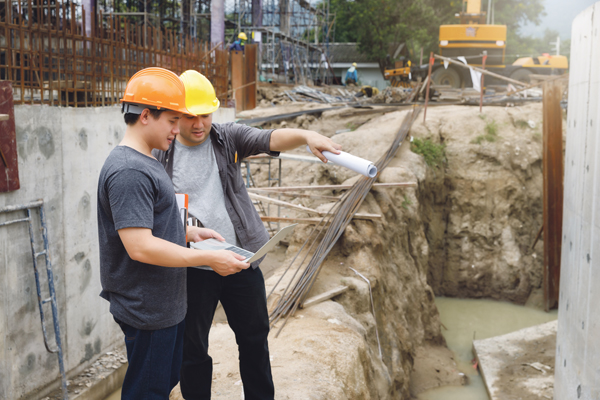 Zwei Handwerker mit Helmen schauen auf einen Plan, hinter ihnen eine Baustelle mit Stahlbeton.